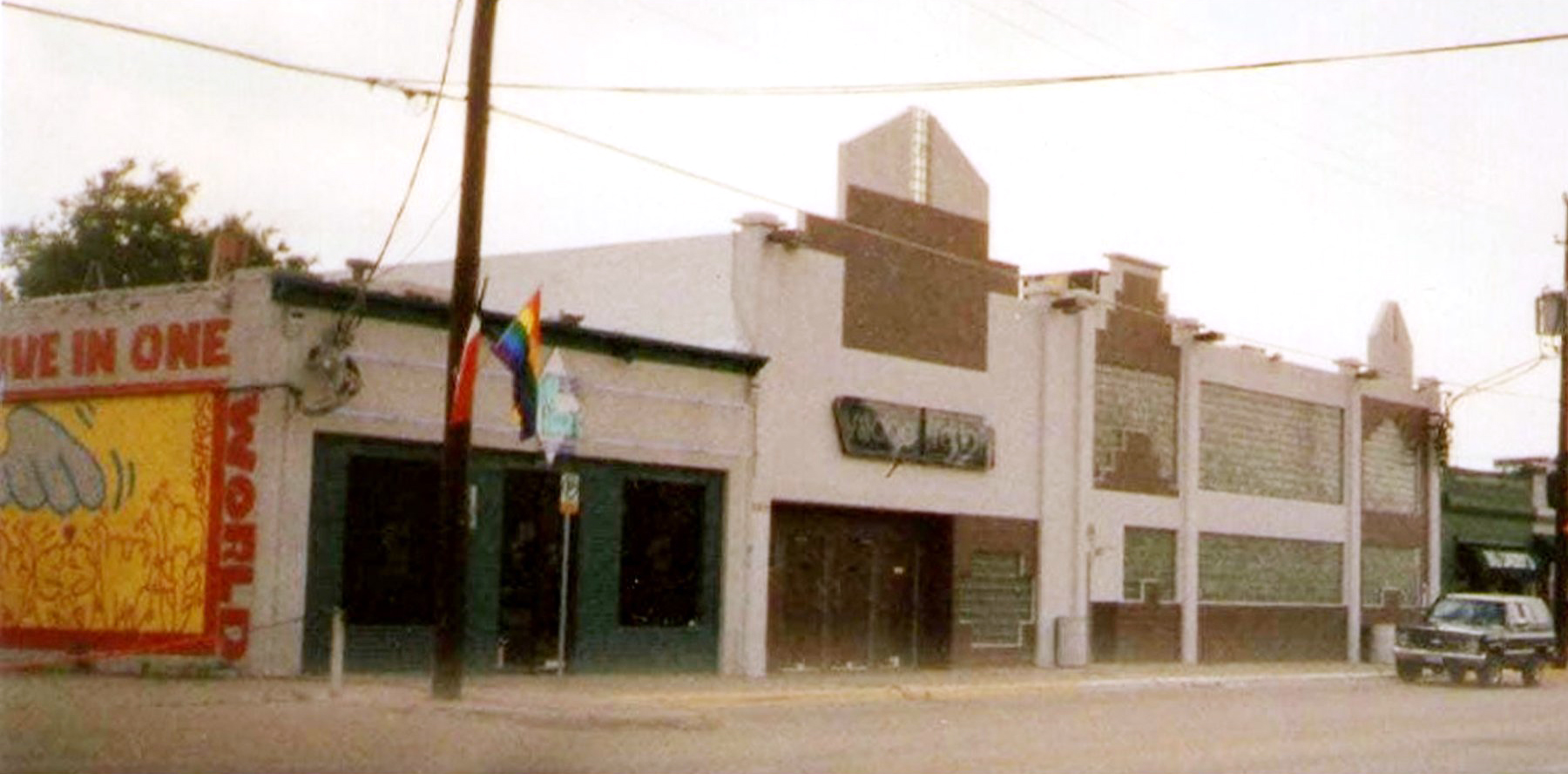 The Old Plantation on Cedar Springs Road, Dallas — a low white building with a rainbow pride flag flying out front and a 'Live in One World' mural on the side. The room Frank Caven opened in the early 1970s.